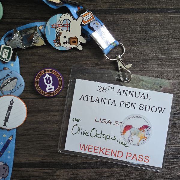 A lanyard full of pins and buttons holding a name tag/weekend pass for the 28th Annual Atlanta Pen Show for Lisa, aka Olive Octopus Ink, and the 2025 Atlanta Pen Show Fountain Pen Day button in purple, yellow, and orange.